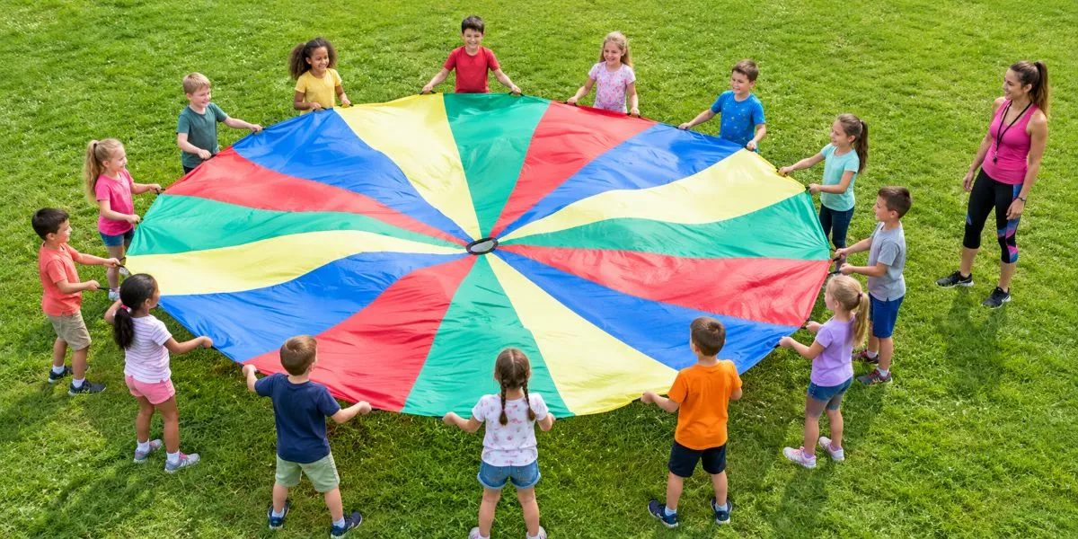 eine nebenberufliche Kindersport Trainerin steht draußen neben einer Gruppe von Kindern, die mit einem bunten Schwungtuch spielen