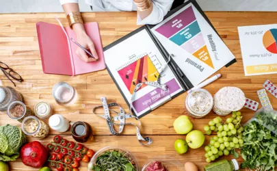 Ernährungsberaterin schreibt Diätplan, Blick von oben auf den Tisch mit verschiedenen gesunden Produkten und Zeichnungen zum Thema gesunde Ernährung