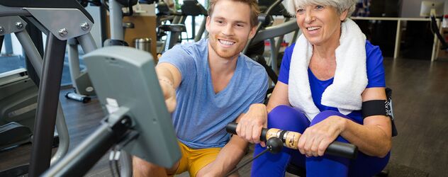 In einem Fitnessstudio ist ein Seniorentrainer gerade dabei eine Seniorin in der Ausübung einer Fitnessübung auf einem der Fitnessgeräte zu unterstützen