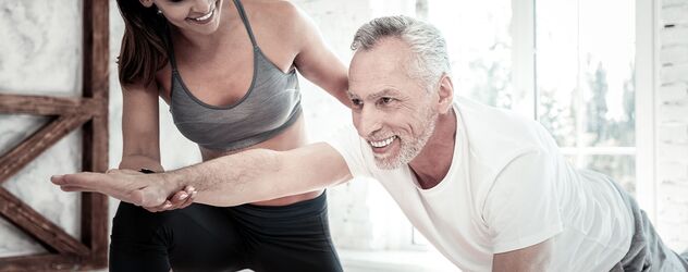 Eine junge Seniorentrainerin ist gerade dabei mit einem ihrer Klienten eine Übung im Rahmen eines Seniorentrainings durchzuführen. Sie sind dabei in einem hell erleuchteten Fitnessstudio