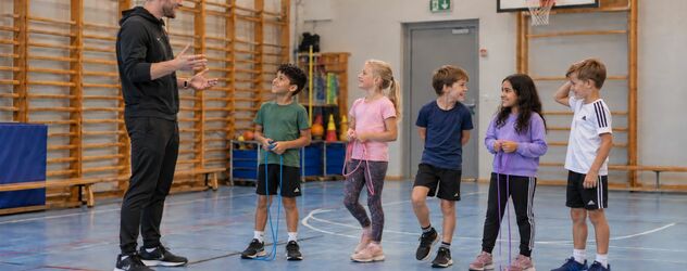 ein Kindersporttrainer erklärt fünf Kindern eine Übung in einer kleinen Sporthalle