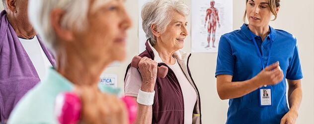 In einem hellen Raum erklärt eine junge Seniorentrainerin mehreren Senioren und Seniorinnen leichte Übungen, die sie mittels kleiner Gewichte ausüben. Dabei stehen sie vor anatomischen Abbildungen des menschlichen Körpers
